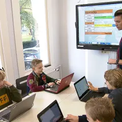 Kinderen leren op laptops in een klaslokaal van een particuliere basisschool. Een leraar staat bij een digibord.