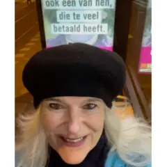 Vrouw met zwarte baret glimlacht naar de camera, met op de achtergrond een poster met de tekst "ook een van hen, die te veel betaald heeft.