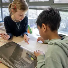Meisje wijst naar tekst in boek, jongen leest mee. Begeleid hardop lezen in de klas.