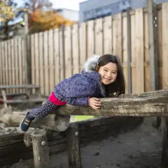 Lachend meisje op een houten speeltoestel in de zandbak van een basisschool. Ze draagt een winterjas en een vrolijke legging.