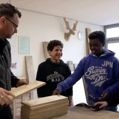 Docent toont hout aan leerlingen. Een van hen, met een blauwe hoodie, lacht. Dit is een cluster 4 indicatie voor praktijkonderwijs.