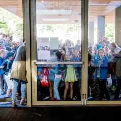 Kinderen staan te dringen bij een glazen deur, nieuwsgierig naar wat er binnen gebeurt. Ze lijken te wachten op iets, misschien wel de uitleg over wat zijn redactiesommen.