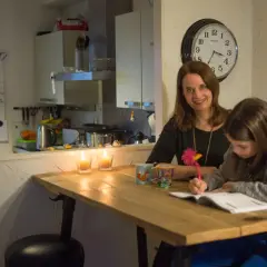 Moeder helpt dochter met huiswerk, terwijl kaarsen branden. Op de muur hangt een klok en een whiteboard met een verhoudingstabel procenten.
