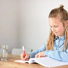 Een meisje schrijft in haar schrift, geconcentreerd op het oplossen van ongelijknamige breuken. Een glas water en fruit staan klaar.