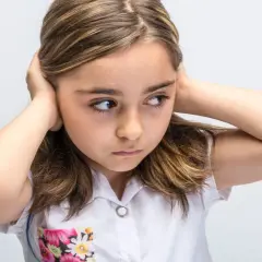 Meisje met handen op de oren, probeert overprikkeling te voorkomen. Ze kijkt weg, alsof ze de wereld even buiten wil sluiten.
