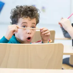 Een jongen van 6 jaar met krullend haar houdt een potlood voor zijn ogen, vol concentratie. Een ander kind steekt een roze potlood omhoog.