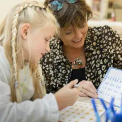 Een juf helpt een meisje met vlechten met leren schrijven groep 1. Ze oefenen letters op een whiteboard.