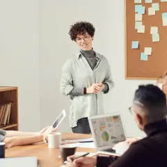 Een vrouw presenteert ideeën voor spreekbeurt onderwerpen aan een team. Post-its op het prikbord suggereren brainstormsessies.