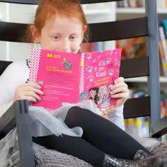 Een meisje met rood haar oefent met lezen in een comfortabele stoel, omringd door boeken.