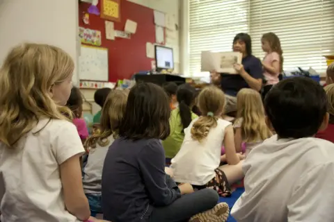 Kinderen zitten aandachtig te luisteren naar de juf die voorleest. Een mooi voorbeeld van leren van elkaar in de klas.