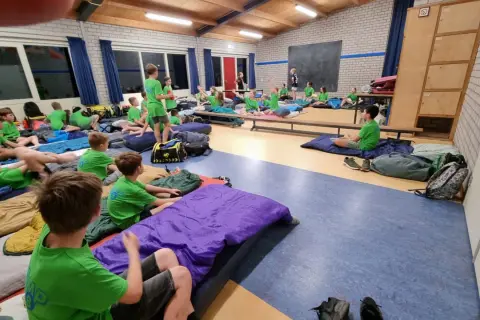 Kinderen van kamp groep 8 liggen op slaapzakken in een gymzaal, klaar voor een nacht vol avontuur.