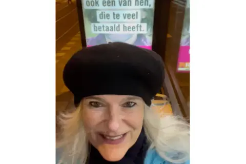 Vrouw met zwarte baret glimlacht naar de camera, met op de achtergrond een poster met de tekst "ook een van hen, die te veel betaald heeft.