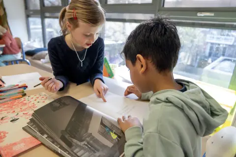 Meisje wijst naar tekst in boek, jongen leest mee. Begeleid hardop lezen in de klas.