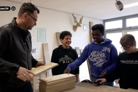 Docent toont hout aan leerlingen. Een van hen, met een blauwe hoodie, lacht. Dit is een cluster 4 indicatie voor praktijkonderwijs.