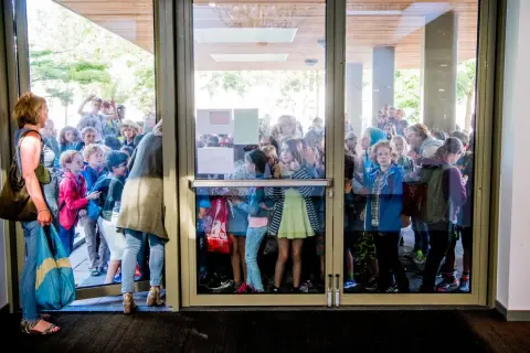 Kinderen staan te dringen bij een glazen deur, nieuwsgierig naar wat er binnen gebeurt. Ze lijken te wachten op iets, misschien wel de uitleg over wat zijn redactiesommen.
