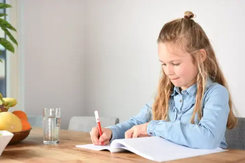 Een meisje schrijft in haar schrift, geconcentreerd op het oplossen van ongelijknamige breuken. Een glas water en fruit staan klaar.