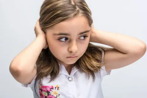 Meisje met handen op de oren, probeert overprikkeling te voorkomen. Ze kijkt weg, alsof ze de wereld even buiten wil sluiten.