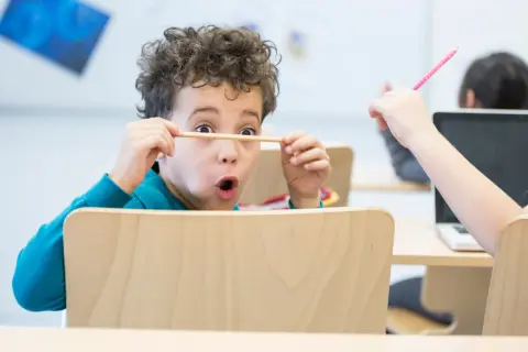 Een jongen van 6 jaar met krullend haar houdt een potlood voor zijn ogen, vol concentratie. Een ander kind steekt een roze potlood omhoog.