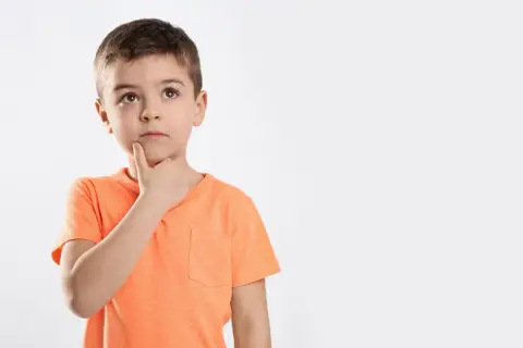 Jongetje met oranje shirt denkt diep na, misschien een beetje traag van begrip, met zijn vinger op zijn kin.