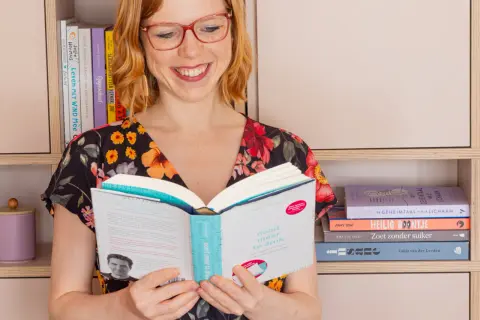 Vrouw met bril leest boek, "nooit meer te druk". Ze is omringd door boeken, wat een **betrekkelijk voornaamwoord** is voor haar passie.