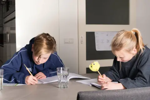 Twee kinderen maken huiswerk aan tafel. De jongen schrijft met een potlood, de meid met een leuke gele pen. Ze oefenen met delen met kommagetallen.