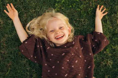 Lachend meisje met blonde krullen ligt op gras. Ze geniet van de rust, een moment van kalmte bij onderprikkeling ADHD.