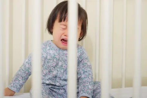 Een huilend kind in een ledikant, met gesloten ogen en open mond. Het kind draagt een pyjama met bloemenprint. Dit kan duiden op sensorische overprikkeling, zoals bij hoofd bonken autisme.