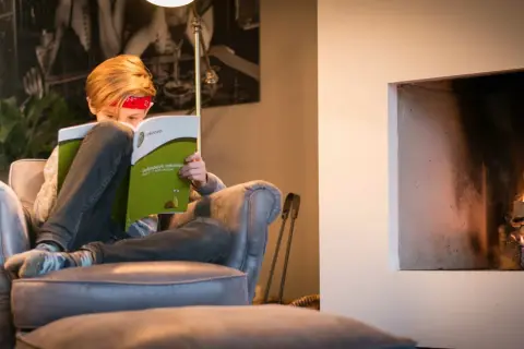Kind met rood bandje leest een oefenboek rekenen bij de haard.