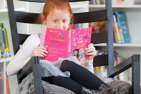 Een meisje met rood haar oefent met lezen in een comfortabele stoel, omringd door boeken.