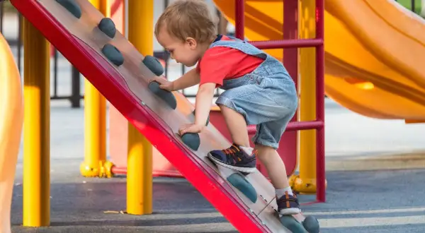 Een peuter klimt op een speeltoestel. Dit is een voorbeeld van hoe kinderen spelenderwijs leren, wat betekent bso: buiten school opvang, waar ze dit soort activiteiten kunnen doen.