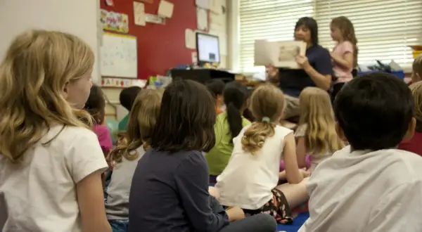 Kinderen zitten aandachtig te luisteren naar de juf die voorleest. Een mooi voorbeeld van leren van elkaar in de klas.
