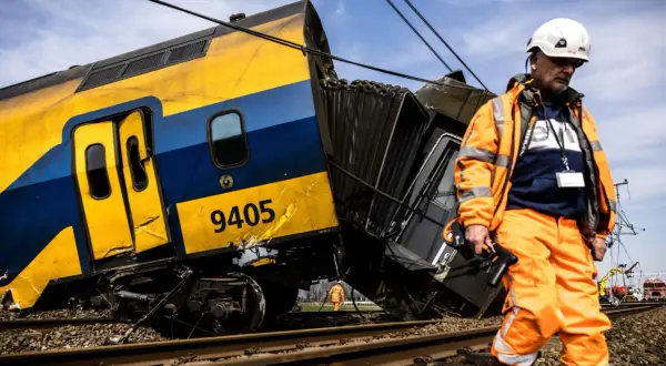 Een ontspoorde trein, nummer 9405, ligt scheef op het spoor. Een man in oranje werkkleding inspecteert de schade, de wagons lijken per ongeluk aan elkaar of los te zijn geraakt.