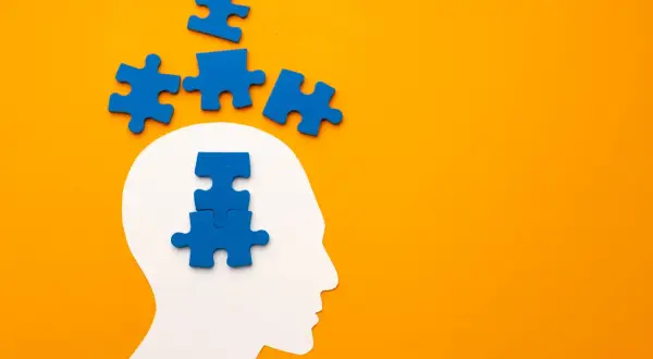 Blauwe puzzelstukjes vallen uit een witte hoofdvorm, sommigen boven, anderen in het hoofd. Het symboliseert het proces van het oplossen van gedachten, zoals bij pdd nos engels.
