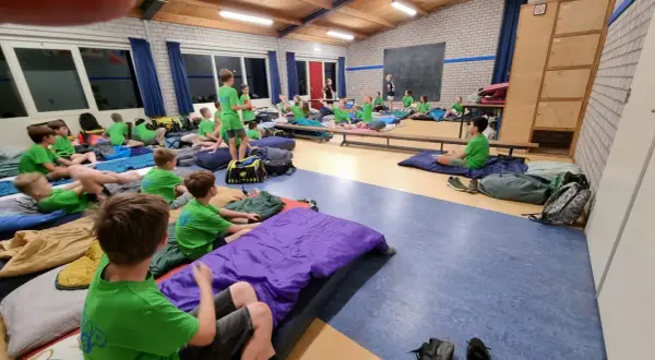 Kinderen van kamp groep 8 liggen op slaapzakken in een gymzaal, klaar voor een nacht vol avontuur.