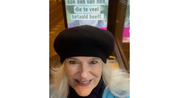 Vrouw met zwarte baret glimlacht naar de camera, met op de achtergrond een poster met de tekst "ook een van hen, die te veel betaald heeft.
