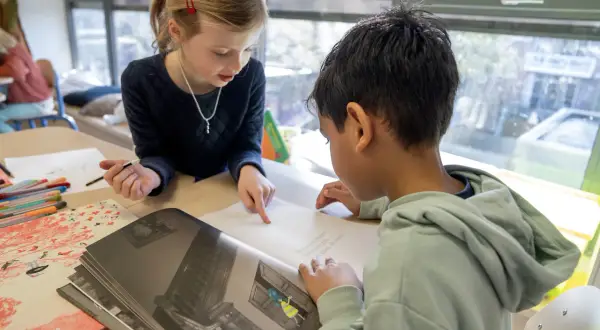 Meisje wijst naar tekst in boek, jongen leest mee. Begeleid hardop lezen in de klas.