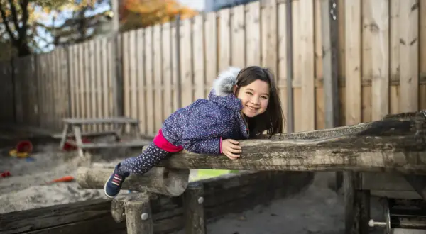 Lachend meisje op een houten speeltoestel in de zandbak van een basisschool. Ze draagt een winterjas en een vrolijke legging.