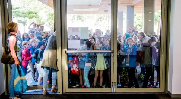 Kinderen staan te dringen bij een glazen deur, nieuwsgierig naar wat er binnen gebeurt. Ze lijken te wachten op iets, misschien wel de uitleg over wat zijn redactiesommen.