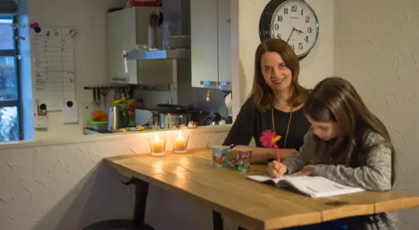 Moeder helpt dochter met huiswerk, terwijl kaarsen branden. Op de muur hangt een klok en een whiteboard met een verhoudingstabel procenten.