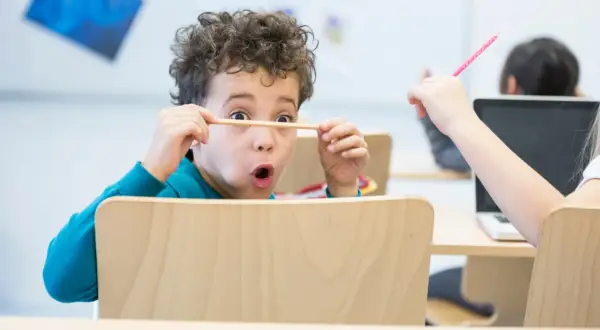 Een jongen van 6 jaar met krullend haar houdt een potlood voor zijn ogen, vol concentratie. Een ander kind steekt een roze potlood omhoog.