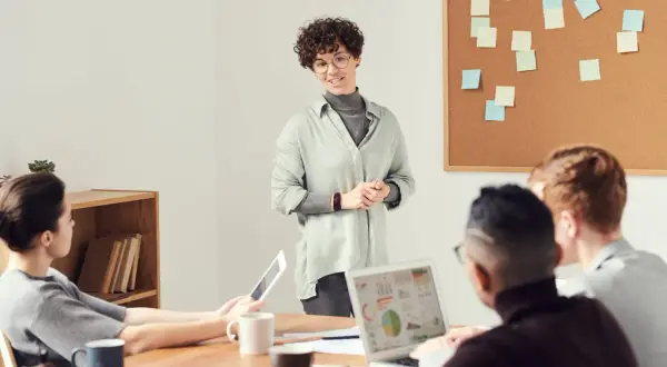 Een vrouw presenteert ideeën voor spreekbeurt onderwerpen aan een team. Post-its op het prikbord suggereren brainstormsessies.
