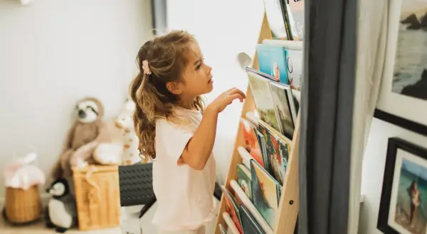Een meisje met blond haar kijkt aandachtig naar de boekenplank, klaar om een nieuw avontuur te ontdekken. Ze houdt van lezen, kinderen.