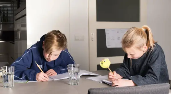 Twee kinderen maken huiswerk aan tafel. De jongen schrijft met een potlood, de meid met een leuke gele pen. Ze oefenen met delen met kommagetallen.