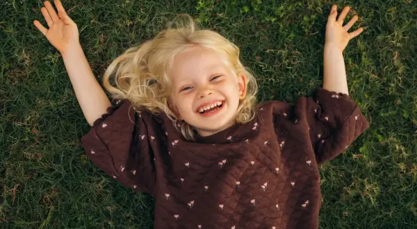 Lachend meisje met blonde krullen ligt op gras. Ze geniet van de rust, een moment van kalmte bij onderprikkeling ADHD.