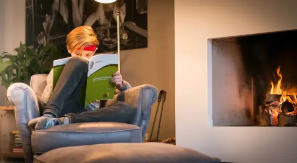 Kind met rood bandje leest een oefenboek rekenen bij de haard.