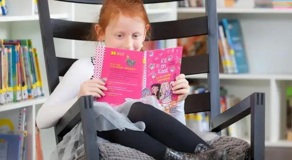 Een meisje met rood haar oefent met lezen in een comfortabele stoel, omringd door boeken.