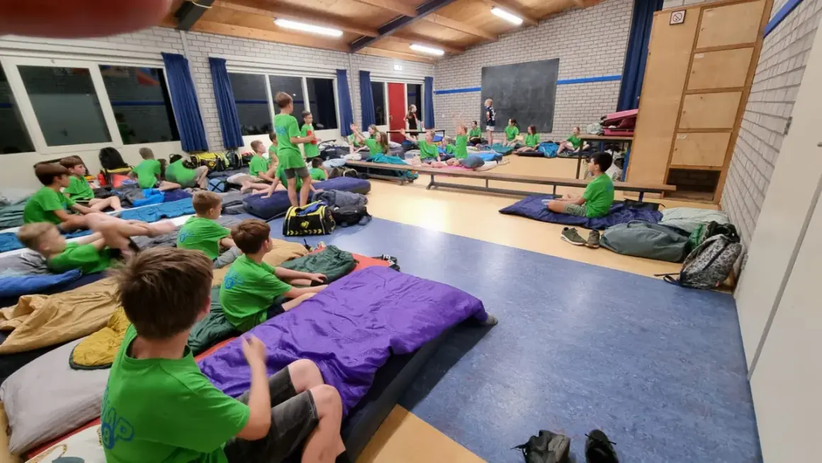 Kinderen van kamp groep 8 liggen op slaapzakken in een gymzaal, klaar voor een nacht vol avontuur.