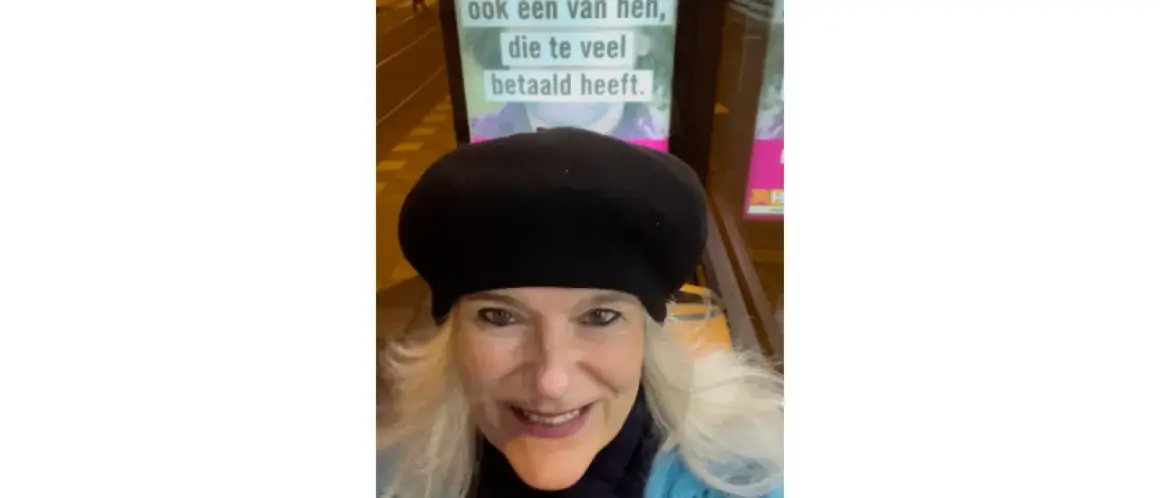Vrouw met zwarte baret glimlacht naar de camera, met op de achtergrond een poster met de tekst "ook een van hen, die te veel betaald heeft.