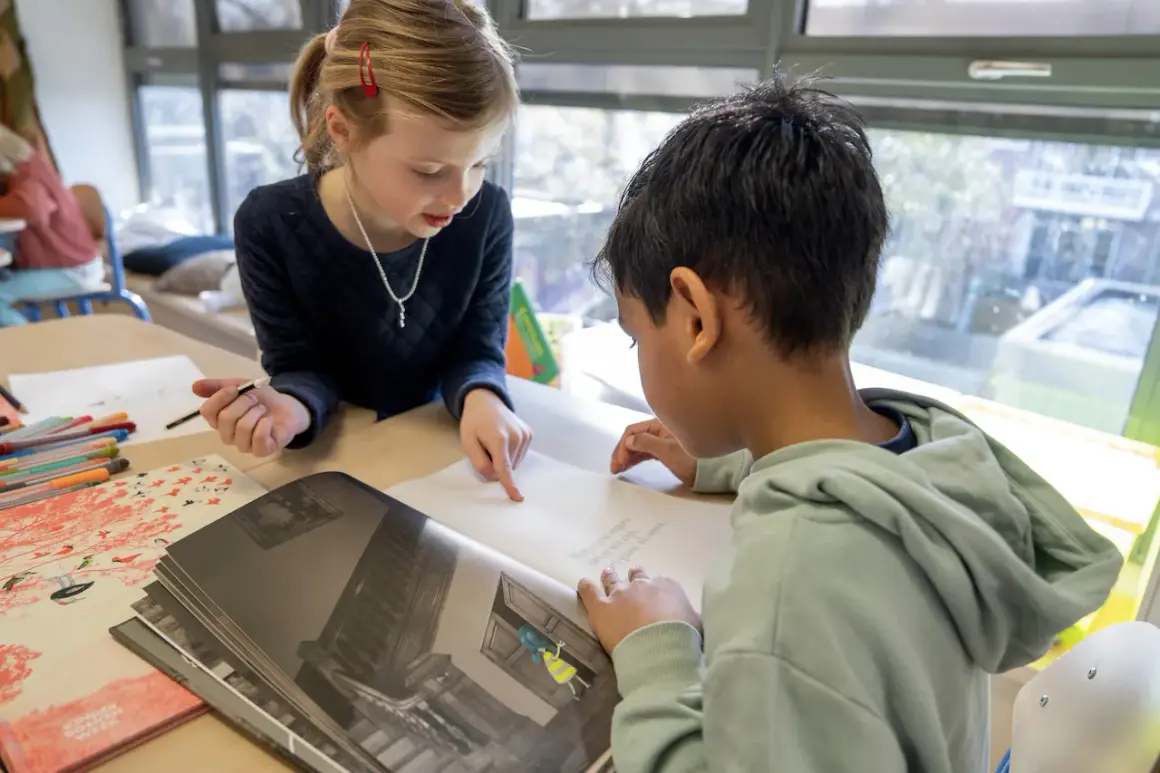 Meisje wijst naar tekst in boek, jongen leest mee. Begeleid hardop lezen in de klas.