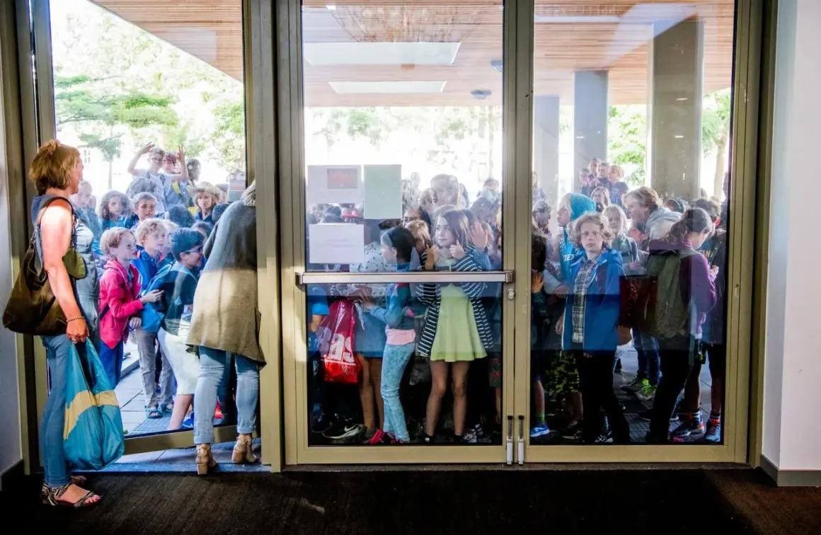 Kinderen staan te dringen bij een glazen deur, nieuwsgierig naar wat er binnen gebeurt. Ze lijken te wachten op iets, misschien wel de uitleg over wat zijn redactiesommen.